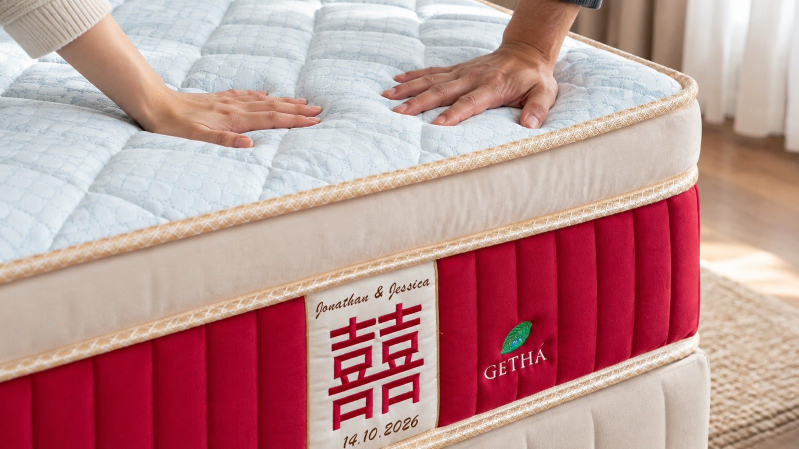 Two hands pressing the pillow top of a Getha Primefibre mattress above the 囍 panel, embroidered with Jonathan & Jessica and 14.10.2026